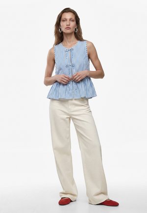 Femme portant un haut sans manches rayé bleu et blanc avec des liens, un pantalon large crème et des chaussures plates rouges, debout sur un fond blanc.