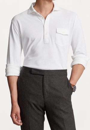 Homme portant un polo blanc à manches longues avec une poche poitrine et des manches retroussées, associé à un pantalon gris foncé et une montre-bracelet.