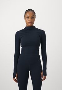 Top nero a maniche lunghe con scollatura alta, realizzato in un tessuto elasticizzato, abbinato a leggings a vita alta coordinati, con texture a coste e branding discreto.