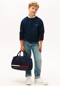 Felpa navy con bordature rosse, jeans azzurri chiari e sneakers bianche. Tiene una borsa a tracolla navy con accenti rossi e bianchi, con logo.