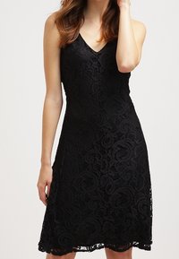 Robe en dentelle noire avec un décolleté en V, sans manches, et un motif floral complexe, se terminant par un ourlet festonné.