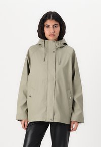 Veste à capuche verte avec une fermeture éclair à l'avant, des boutons-pression, des manches raglan et des poches latérales, fabriquée en tissu lisse et résistant à l'eau.