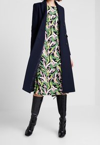 Navy long coat over a multi-colored floral dress with green leaves and pink accents, paired with black knee-high textured boots.