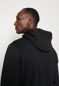 Sudadera con capucha negra de tela suave y texturizada con costuras cosidas. Presenta un ajuste holgado y un diseño de tipo pullover con vista trasera.