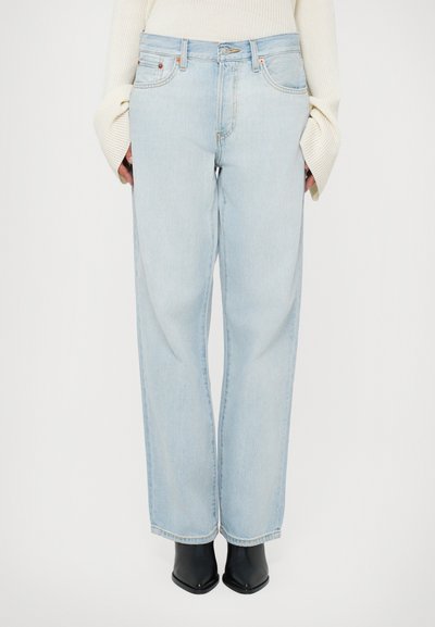 Jean taille haute bleu clair avec une coupe large. Fabriqué en denim, il dispose de poches avant et d'une fermeture éclair. Porté avec des bottines noires.