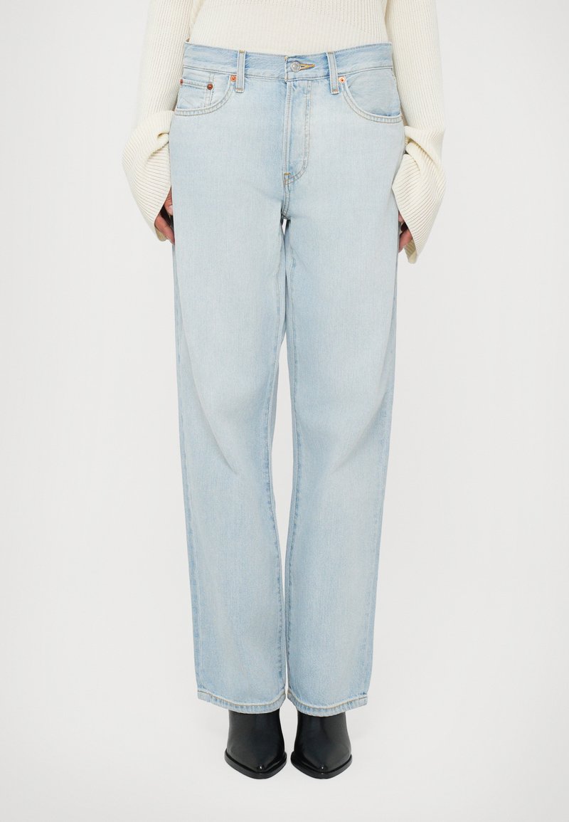 Jeans azzurri chiari a vita alta con una vestibilità ampia. Realizzati in denim, con tasche frontali e chiusura con cerniera. Indossati con stivaletti neri fino alla caviglia.
