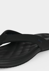 Μαύρο, υφασμάτινο καουτσούκ σαγιονάρι με μαλακό πάτο και διαγώνιο λουράκι που διαθέτει διακριτικό λογότυπο «Reef» στο πλάι του λουριού.