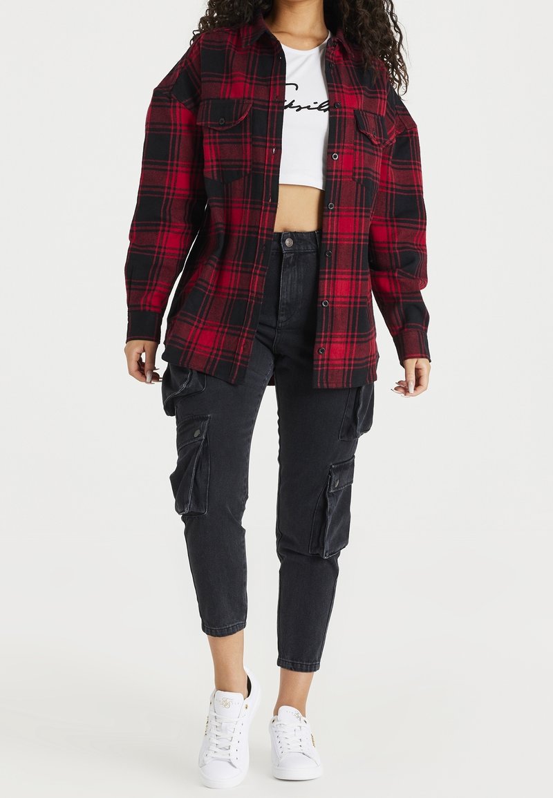 Une chemise en flanelle à carreaux rouge et noir avec deux poches avant, portée par-dessus un haut court blanc, associée à un pantalon cargo noir et des baskets blanches.