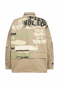 Beige Oversize-Jacke mit grafischen Drucken in Schwarz und Creme, grünen Akzenten und Knopfdetails an den Ärmeln.