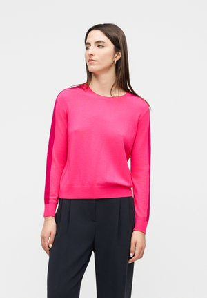 Γυναίκα με μακριά καστανά μαλλιά που φοράει φωτεινό ροζ πουλόβερ με μακριά μανίκια και μαύρο παντελόνι, στέκεται με τα χέρια στα πλάγια, κοιτώντας αριστερά.