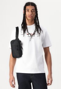 Hvit bomull T-skjorte med grafisk tekst, svart crossbody veske med glidelåser, brukt over mørk denim jeans. Modellen har langt flettehår.