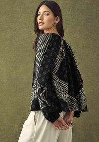 Blouse noire à motifs avec des manches bouffantes, présentant des motifs géométriques blancs. Tissu léger avec une coupe décontractée, associée à un pantalon crème.