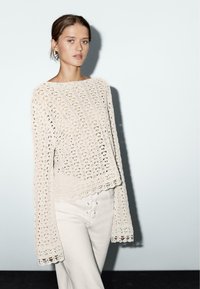 Beige gestrickter Pullover mit weiten Ärmeln, der ein offenes Maschenmuster und gewellte Kanten aufweist. Kombiniert mit weißen Hosen, die über eine Schnürdetails verfügen.