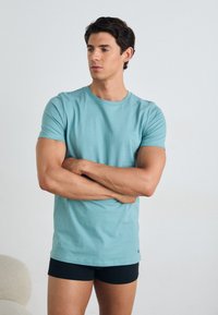 Light blue cotton T-shirt with short sleeves, crew neck. Paired with black shorts, model stands with arms crossed, displaying a fitted silhouette.