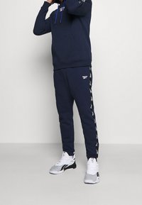 Tmavě modrá mikina s kapucí a přední kapsou, doplněná tmavě modrými tepláky s bílou boční páskou a logem Reebok na stehně.