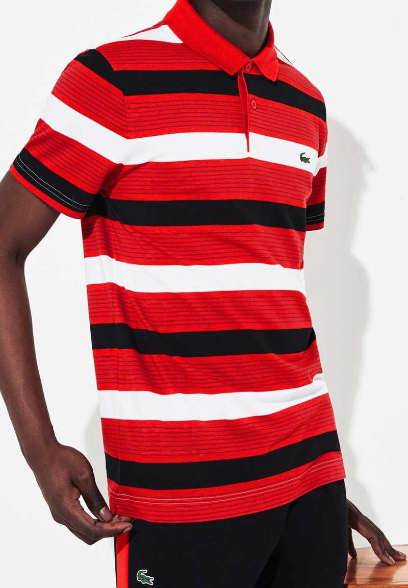 Homme portant un polo à rayures rouges, blanches et noires et un pantalon noir, debout avec une main tenant l'ourlet du polo.