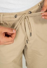 Los pantalones cortos beige cuentan con una cintura elástica con cordones ajustables, abrazaderas metálicas en contraste y una tela ligera. Se están ajustando los cordones.