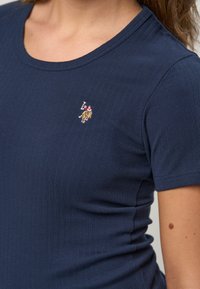 Nærbillede af en person iført en marineblå, ribstrikket T-shirt med korte ærmer og broderet polospillerlogo på venstre bryst.