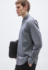 Πουκάμισο με γκρι κουμπιά και διακριτικό καρό μοτίβο, μακριά μανίκια και γιακά, φορεμένο με μαύρη τσάντα, πάνω σε ανοιχτό φόντο.