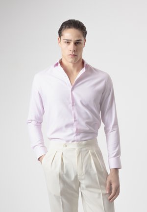 Joven con el pelo peinado hacia atrás, vestido con una camisa rosa claro y pantalones blancos plisados de cintura alta, de pie frente a un fondo liso.
