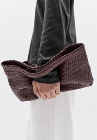 Vævet burgunderlædertaske med en afslappet design og en enkelt hank, vist holdt mod en ensfarvet baggrund.