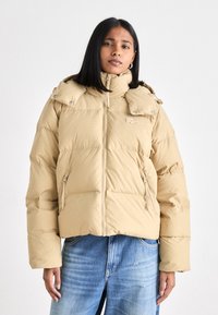 Giacca a vento beige con colletto alto, tasche con zip e bottoni a pressione. Presenta un design trapuntato ed è abbinata a jeans in denim blu.