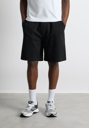 Person iført sorte knælange shorts, hvide crew-strømper, hvide og grønne løbesko, stående med hænderne i lommerne.