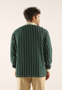 Mand med krøllet hår iført en mørkegrøn langærmet skjorte med hvide lodrette striber og beige bukser, vendt væk fra kameraet.