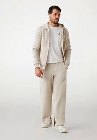 Beige hoodie met witte strepen op de mouwen, gecombineerd met bijpassende beige broek met zijsplitten. Witte T-shirt en sneakers maken de outfit compleet.