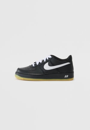 Sort low-top Nike Air sneaker med hvid swoosh-logo, hvide snørebånd og gummi-farvet sål på en ensfarvet lysegrå baggrund.