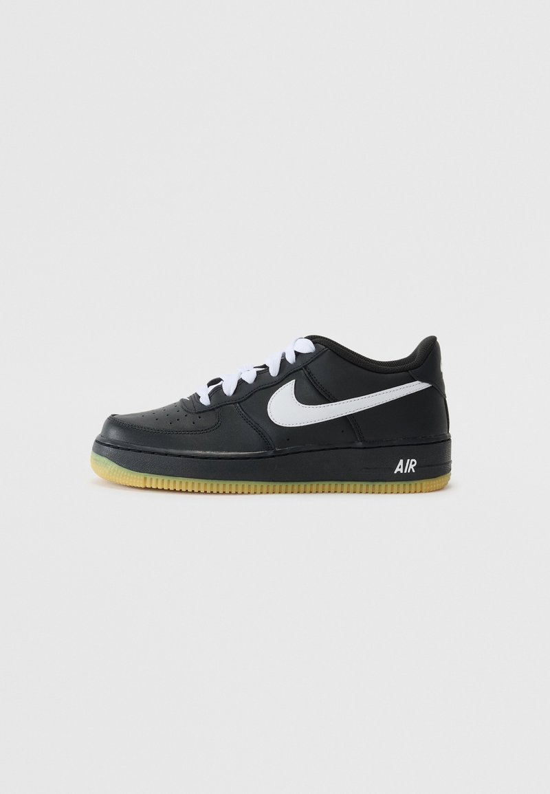 Basket Nike Air noire basse avec logo Swoosh blanc, lacets blancs et semelle couleur gomme sur fond gris clair uni.