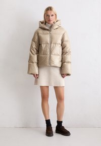 Beżowa puchowa kurtka z kapturem, z zamkiem błyskawicznym, szerokimi rękawami i błyszczącym wykończeniem, połączona z jasną spódnicą i ciemnymi butami.