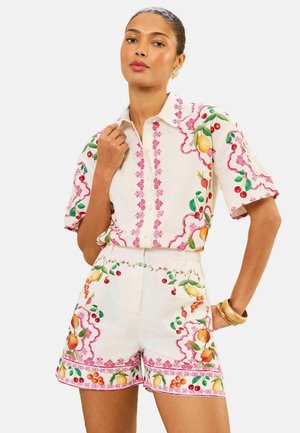 Woman wearing white short-sleeve button-up shirt and matching shorts with fruit and pink floral patterns, accessorized with gold bracelets.