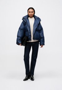 Námořnická puffer bunda, lesklý materiál, oversize střih, velký límec, nošena přes béžový svetr, v kombinaci s tmavými džínami a černou kabelkou.