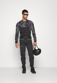 Équipement de plein air pour hommes avec un haut à manches longues à motif noir et un pantalon noir. Comprend des fermetures éclair, des poches et un casque noir tenu à la main.