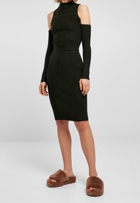 Svart ribbad stickad klänning med en hög krage och utskärningar på axlarna, ihopparad med bruna fluffiga slidesandaler.