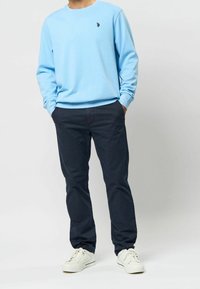 Vaaleansininen collegepaita pitkillä hihoilla ja pyöreällä kaula-aukon reunuksella, yhdistetty tummansinisiin housuihin ja valkoisiin lenkkareihin. Paidassa pieni musta logo.