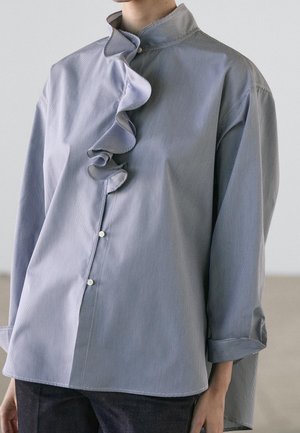 Person wearing a light blue button-up shirt with vertical stripes and a ruffled detail running along the collar and front placket.