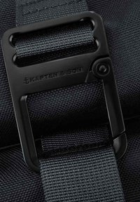 Svart remspänne från Kapten & Son, med en texturerad yta, graverad logotyp och en säker klämehanism. Hållbart syntetiskt material.