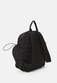 Mochila de tela negra con textura suave, cierre de cordón, correas ajustables y un asa en la parte superior. Cuenta con un bolsillo lateral con detalles metálicos.