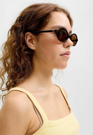 Młoda kobieta z kręconymi brązowymi włosami, nosząca czarne, okrągłe okulary przeciwsłoneczne i jasnżółtą, bezrękawnikową bluzkę, zwrócona w prawo na jednolitym tle.