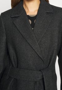 Cappotto in misto lana grigio scuro con texture a spina di pesce, revers, vita con cintura e maniche lunghe. Motivo sottile visibile sotto.