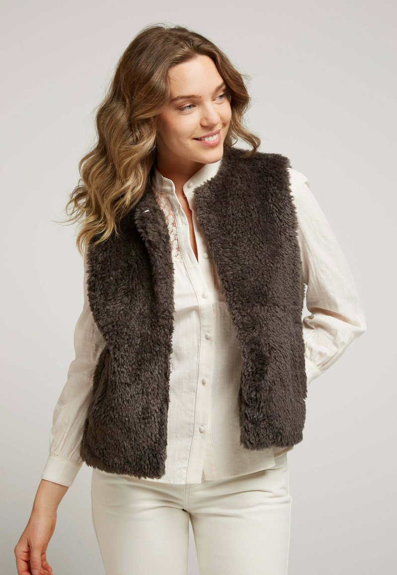 Gilet en fausse fourrure de couleur gris foncé, avec un col haut et un design ouvert à l'avant. Porté sur une blouse de couleur claire et associé à un pantalon crème.