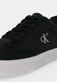 Baskets en cuir noir avec logo blanc "cK" sur le côté et semelle épaisse en caoutchouc blanc, avec lacets noirs et couture détaillée.