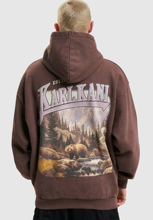 Personne aux cheveux blonds courts portant un sweat à capuche marron avec un graphique de forêt et d’ours et le texte « KARL KANI » dans le dos.