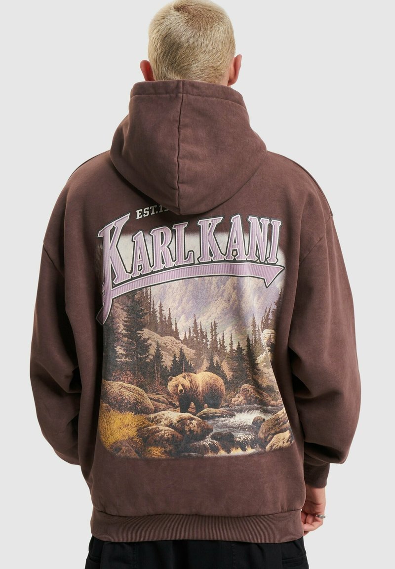 Personne aux cheveux blonds courts portant un sweat à capuche marron avec un graphique de forêt et d’ours et le texte « KARL KANI » dans le dos.