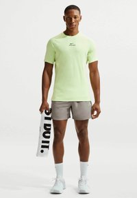 Αθλητικός άνδρας στέκεται κρατώντας λευκή πετσέτα με το κείμενο "JUST DO IT.", φοράει ανοιχτό πράσινο μπλουζάκι Nike Pro, γκρι σορτς, λευκές κάλτσες και λευκά αθλητικά παπούτσια.