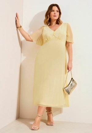 Frau in einem blassgelben Midikleid mit durchsichtigen, flatternden Ärmeln, die eine metallic-goldene Handtasche hält und goldene Absatzschuhe mit Knöchelriemen trägt, steht vor einer weißen Wand.