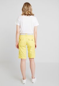 Shorts jaunes avec une ceinture, deux poches arrière et un ourlet replié. Portés avec un t-shirt blanc uni et des chaussures blanches sans lacets.