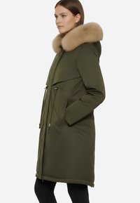 Parka verde oliva con ampio colletto in pelliccia sintetica, chiusura a zip, vita regolabile con cordoncino e due tasche laterali. Tessuto liscio e resistente all'acqua.